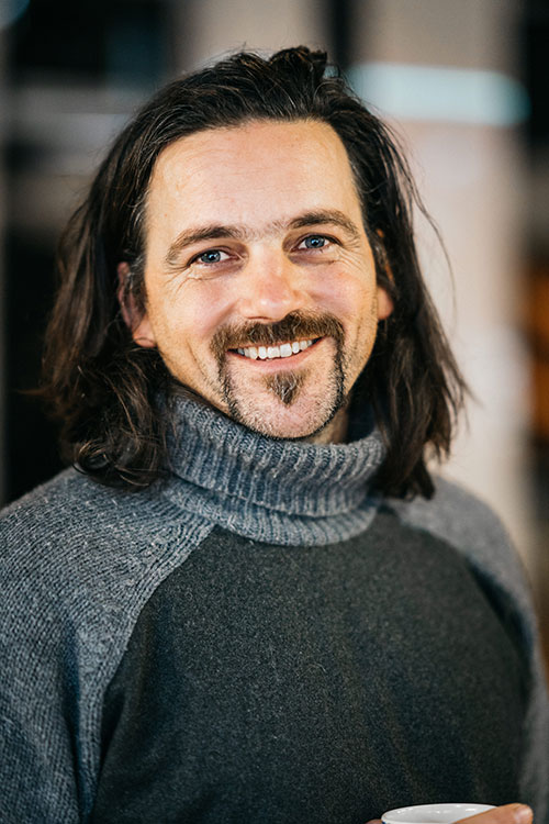 Thor - a medium close-up of a white man with dark hair and a goatee. He wears a grey turtleneck and is smiling.