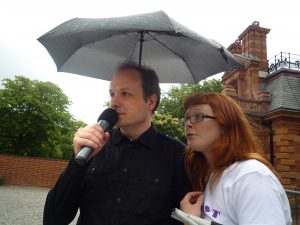 A white male talking into microphone, accompanied by a white woman. They are standing underneath an umbrella.