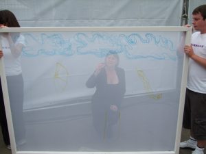 Two people hold a large semi transparent screen. The outline of a woman can be seen on the other side, where she is crouching and drawing.