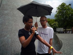 A black woman and white man under an umbrella with a big cone in the background.