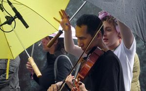 An East Asian man playing viola with audience around him and umbrella over him