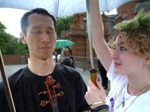 An East Asian man holding his viola under an umbrella held up by his access worker.