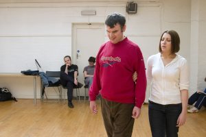 One actor leads another on stage while two other actors sit on chairs off stage