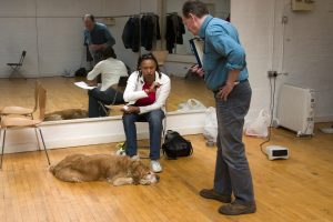 Richard holding a clip board leans over talking to Maria sitting, all reflected in a studio mirror behind