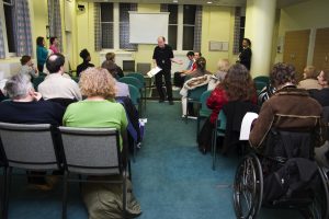 Two banks of audience members at the RNIB with Director centre stage talking to them and behind him, two cast members on stage