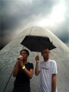 A low angle shot of a black woman speaking into a microphone next to a white man holding an umbrella over their heads, with a giant cone in the background.