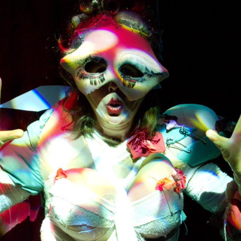 A closeup of a burlesque performer with her yellow gloved hands up, fingers spread. She is wearing a distorted red and white patterned eye mask and crisscrossing bandages across her torso.