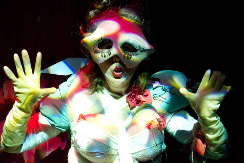 A closeup of a burlesque performer with her yellow gloved hands up, fingers spread. She is wearing a distorted red and white patterned eye mask and crisscrossing bandages across her torso.