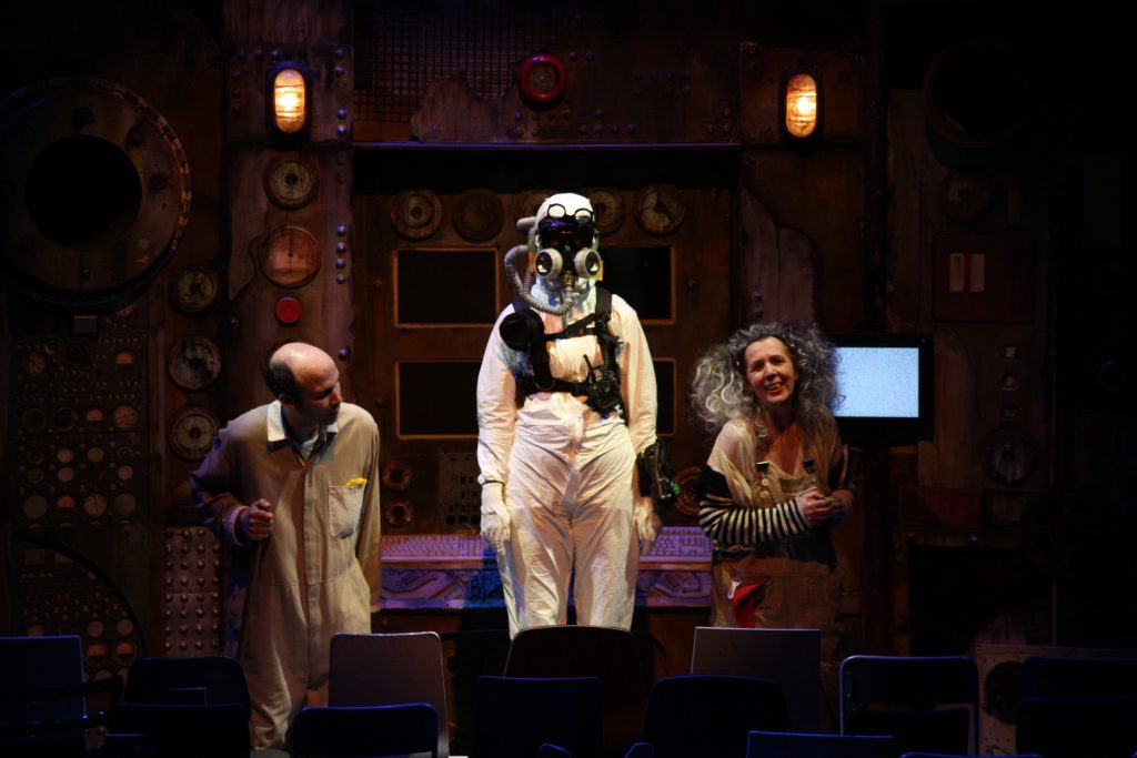 A trio of performers on stage. From left to right, a bald white man wearing a beige jumpsuit with his neck craned to the middle, a person wearing a full body white hazmat suit standing elevated, and an older woman with curly grey hair smiling with hands clutched.