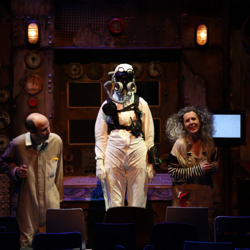 A trio of performers on stage. From left to right, a bald white man wearing a beige jumpsuit with his neck craned to the middle, a person wearing a full body white hazmat suit standing elevated, and an older woman with curly grey hair smiling with hands clutched.