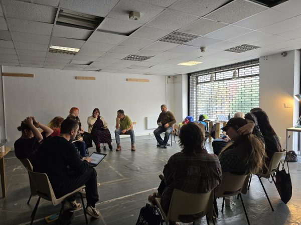 A group of 12 people are sat round on chairs in a circle chatting in a darkly lit space. There’s a large square window to the right.