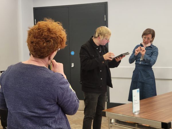 Three people are participating in Touch Tour Training. The facilitator, a man, stands in the middle between two women, looking at an Ipad. They are engaging in conversation.