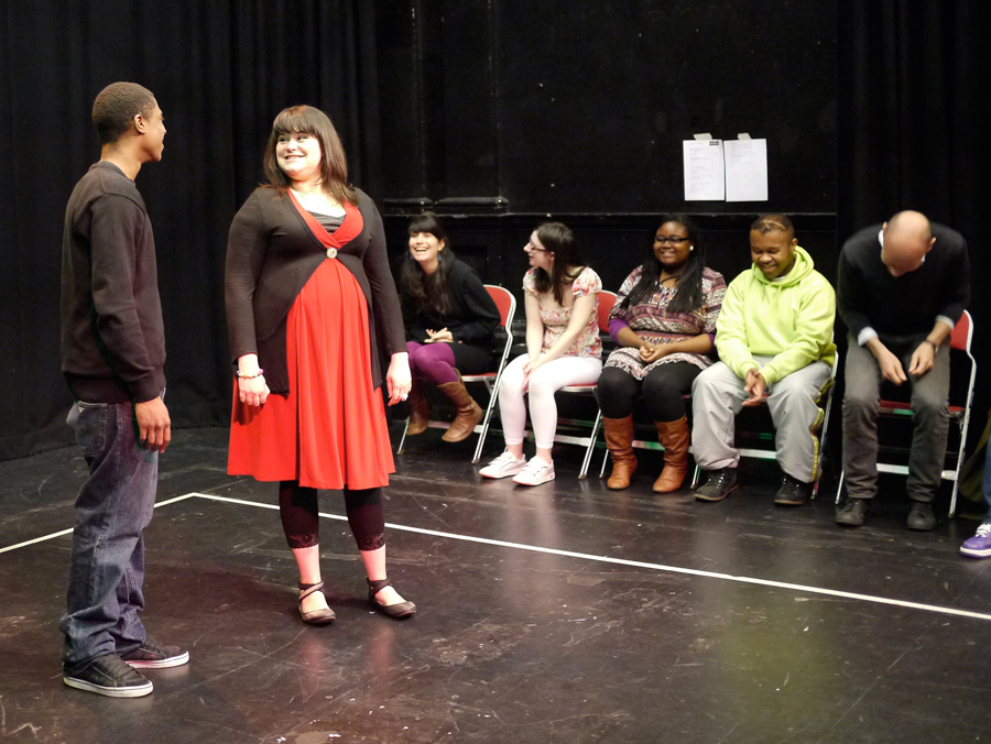 A black male Forum Assistant and a young white female actor perform side by side, smiling in conversation. Five other actors, black and white, look on as audience laughing at them.