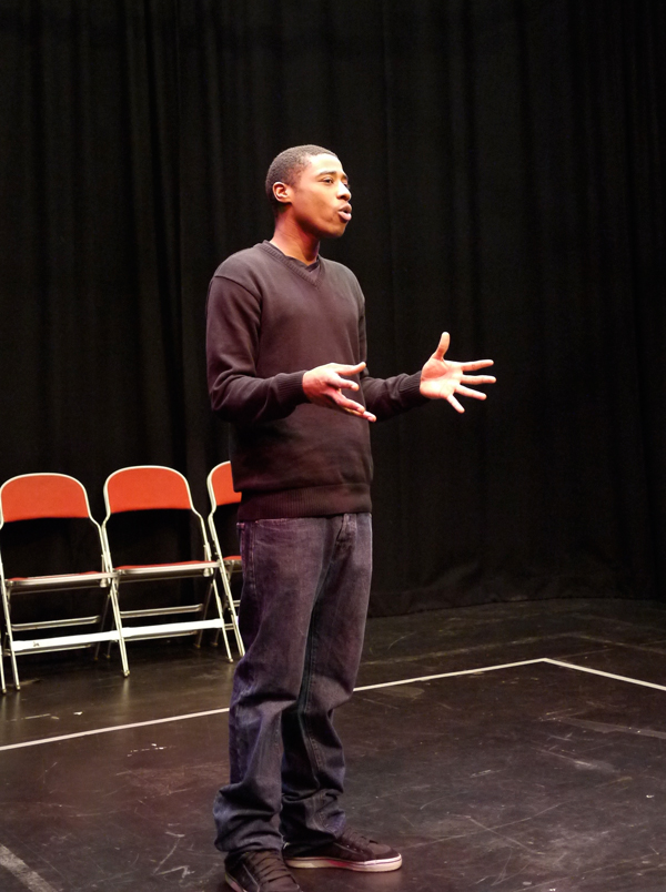 A black Forum Assistant stands centrally, fingers splayed expressively as he explains something to the group, who aren’t visible in the shot.