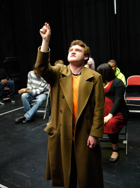 A young white actor playing the role of ‘Teacher’ stands in a long gold coloured overcoat, looking up and reaching for an imaginary object with his right hand.