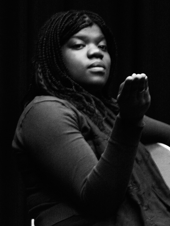 A black and white close up of a black female actor; seated, right hand held up in line with her face. She stares intently at her hand as it appears to unfold.