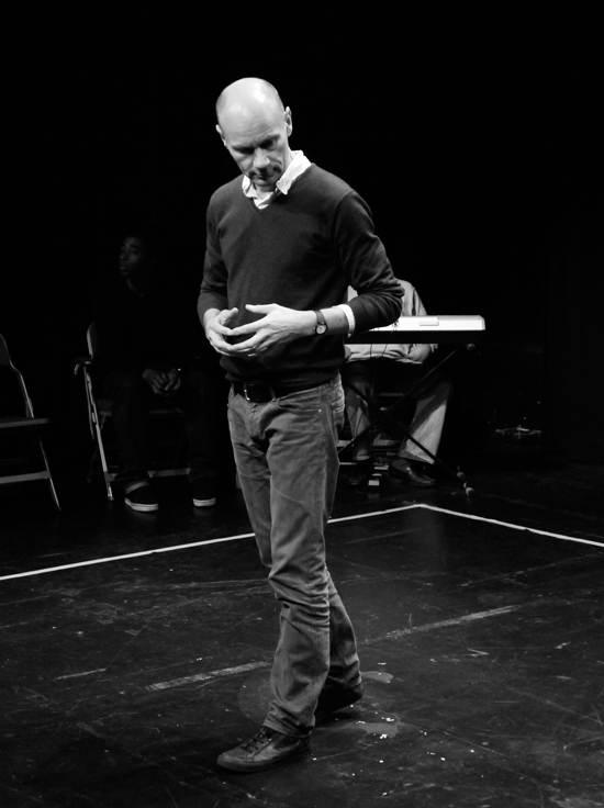 A black and white shot. The director stands alone centre stage, contemplative, hands resting by his stomach and fingers pressed together. His head twists downwards in thought.