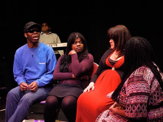 Three black actors and one white actor, all male and female, sit together having a conversation. Another actor is in the background relaxing in front of a keyboard.