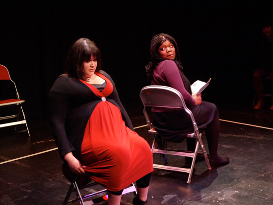 Two female actors, one white and one black, sit on an imaginary bus, one facing front and the other to the side reading a book. One actor twists round to look at the other, a questioning look on her face and pursed lips.