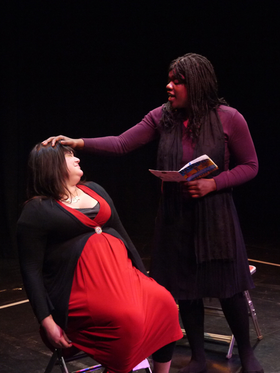 The black female actor stands reading aloud from a book whilst holding her hand over the head of the seated white female actor, who squirms uncomfortably.