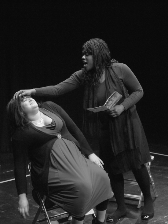 A black and white photo. The black female actor stands reading aloud from a book whilst holding her hand over the head of the seated white female actor, who squirms uncomfortably.