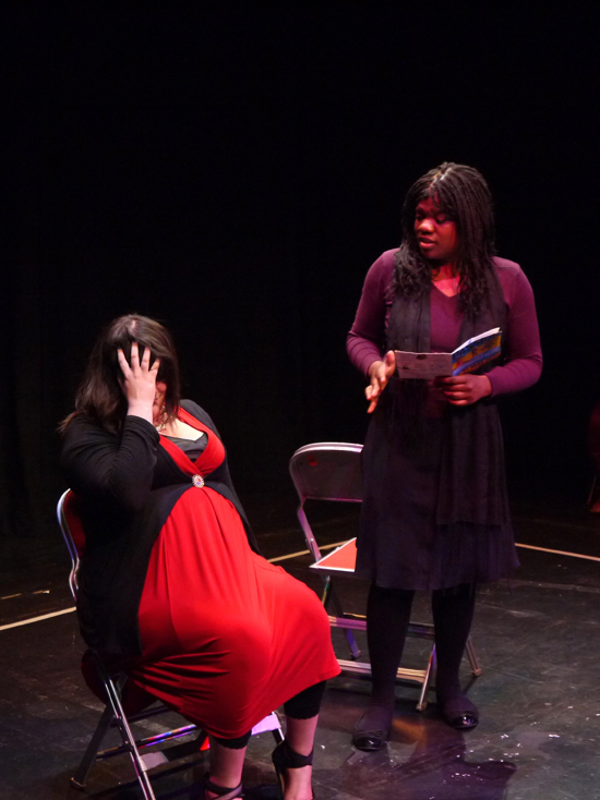 The young white actress sits on the imaginary bus with her head in her hand. The black female actor stands talking to her; she is holding an open book.