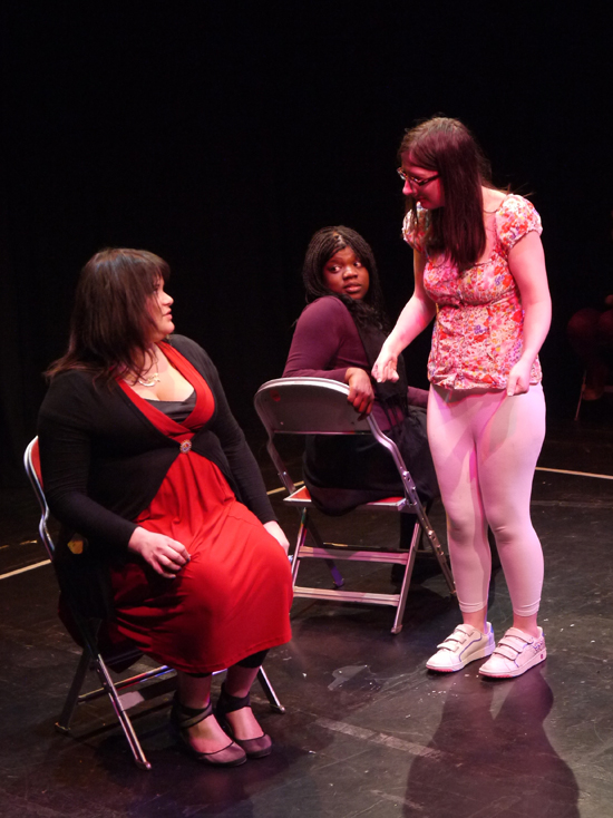 On the bus, a white actor with an imaginary pram approaches the young female white actor, who looks taken aback. The black female actor with the book looks round inquisitively. 