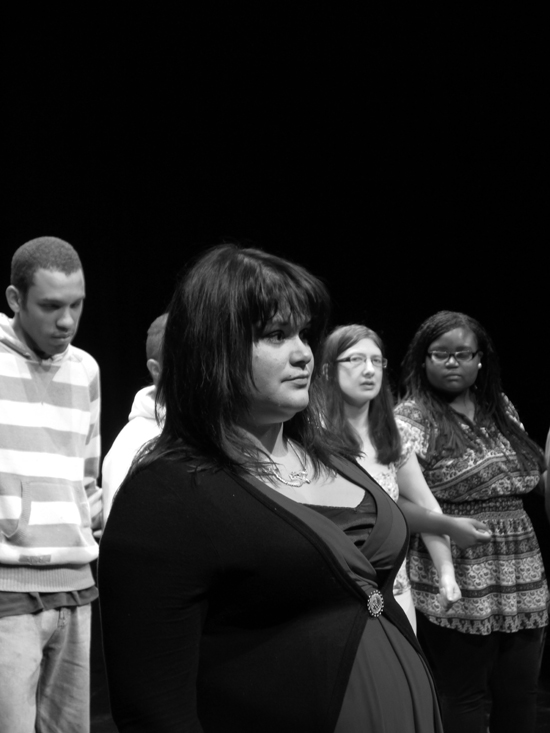 A black and white close up of a young white female actor staring straight ahead, a line of four actors can be seen in the background.