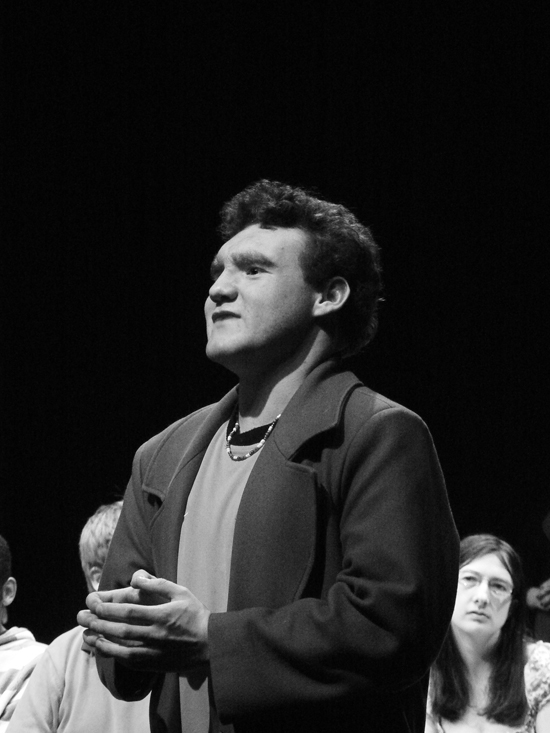 A close up of the young male white actor playing ‘Teacher’. As he talks to the group, his hands are resting at his stomach with his fingers pressed lightly together. Photo in black and white.