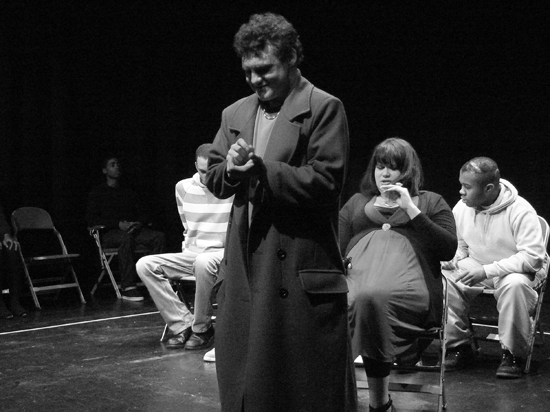 The male actor playing ‘Teacher’ stands with a grin, looking at his hands which appear to hold something near his chest. Three actors sit behind him, distracted by other things.