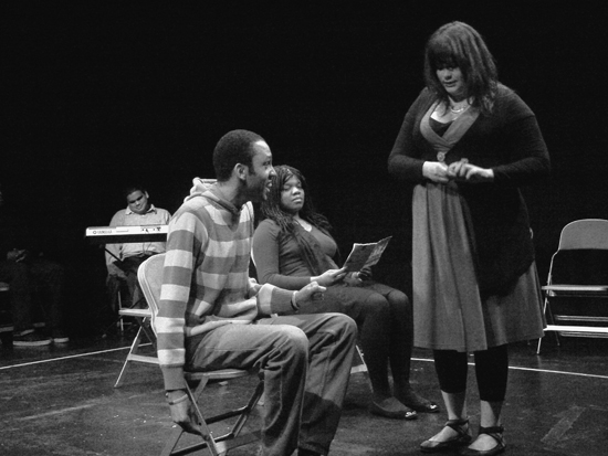 The young white female actor stands on the imaginary bus; a black male actor is seated talking to her as she plays with her fingers uncomfortably. The black female actor with the book looks on. Photo in black and white.