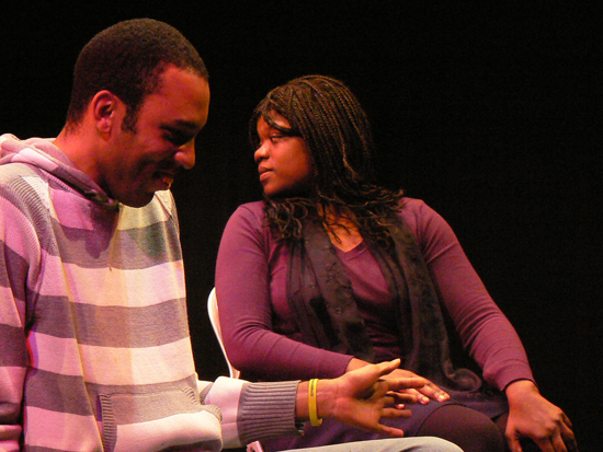 A close up of two black male and female actors, one smiling ahead, the other looking behind her.