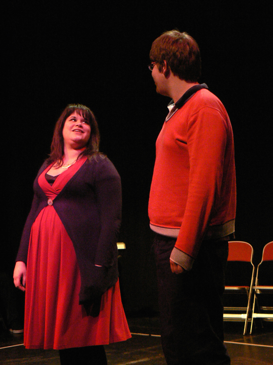 Two white actors male and female at an imaginary cinema; they stand smiling at each other in conversation.