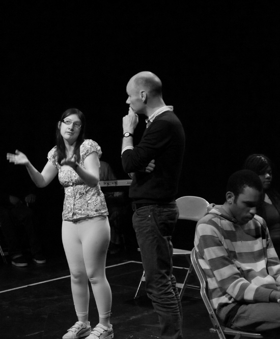 The director talks to a young white female actor who is expressing herself with hand gestures. Photo in black and white.