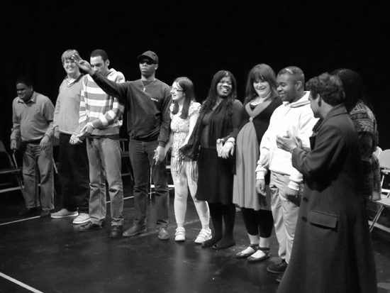 A shot of the whole group of actors in a line and smiling as if preparing for a final bow. Photo in black and white