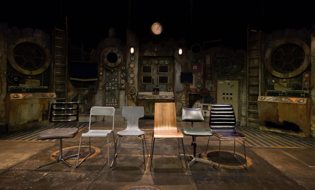 Six chairs lined up on stage in a variety of styles and shapes: some metal and wooden, another leathery and plush.