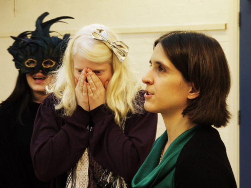 A close up shot of three white females -  The tutor is speaking, another blonde female is holding her hands up to her face, eyes open, and the third is wearing a black and green extravagantly feathered eye mask and laughing.