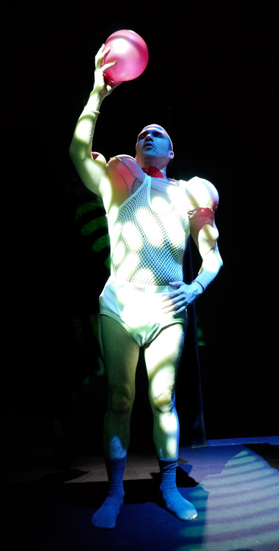 Tim performs as Cataracto in Sheer. Wearing a white fishnet outfit stuffed with inflatables to look like muscles, he stands like a Greek statue, holding up in one hand a bright pink balloon.