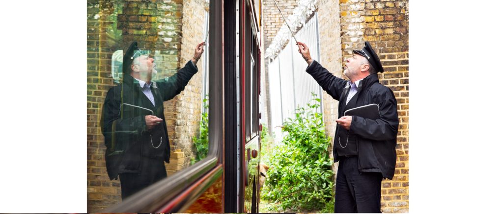 A bus conductor in uniform and cap uses a white cane to point up at the side of a double deck bus