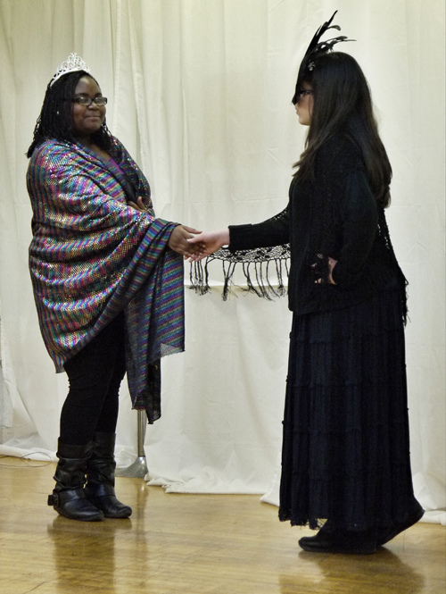The two female actors stand shaking hands in a formal but tentative way. The white actor is wearing long black garments to match the feathered black and green eye mask. The black actor is wearing her long technicoloured shawl and white tiara.