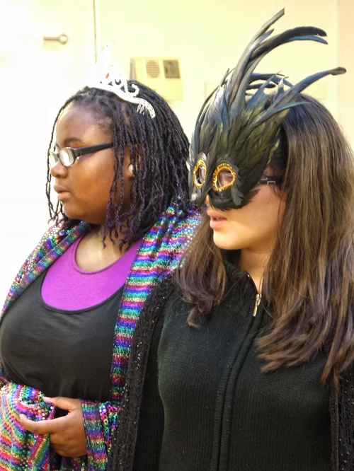Two female actors stand together looking over to the left. The black actor is wearing a long technicoloured shawl and a white tiara and the white actor is wearing a black and green feathered eye mask.