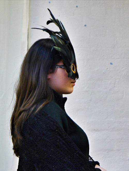 A shot of the female actor with long black garments and black feathered eye mask. She is photographed from head to waist standing in profile.