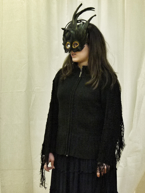 A 3¼ length shot of the female actor posing in her eye mask and black costume against a white background.