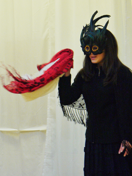 A 3¼ length shot of the female actor with her eye mask and black attire, she is holding up a red towel and saying something.