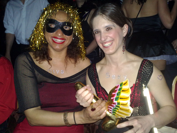 Maria in gold tinsel wig and black mask holding the winged award with Amelia broadly smiling