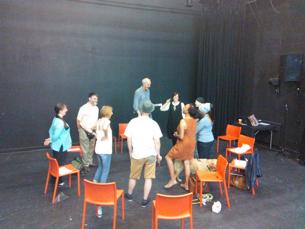 A group of nine people and a guide dog stand in a circle with high energy in a black box theatre