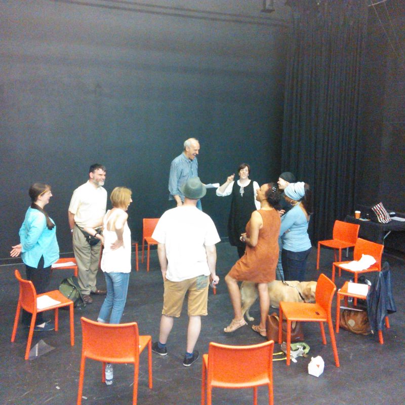 A group of nine people and a guide dog stand in a circle with high energy in a black box theatre