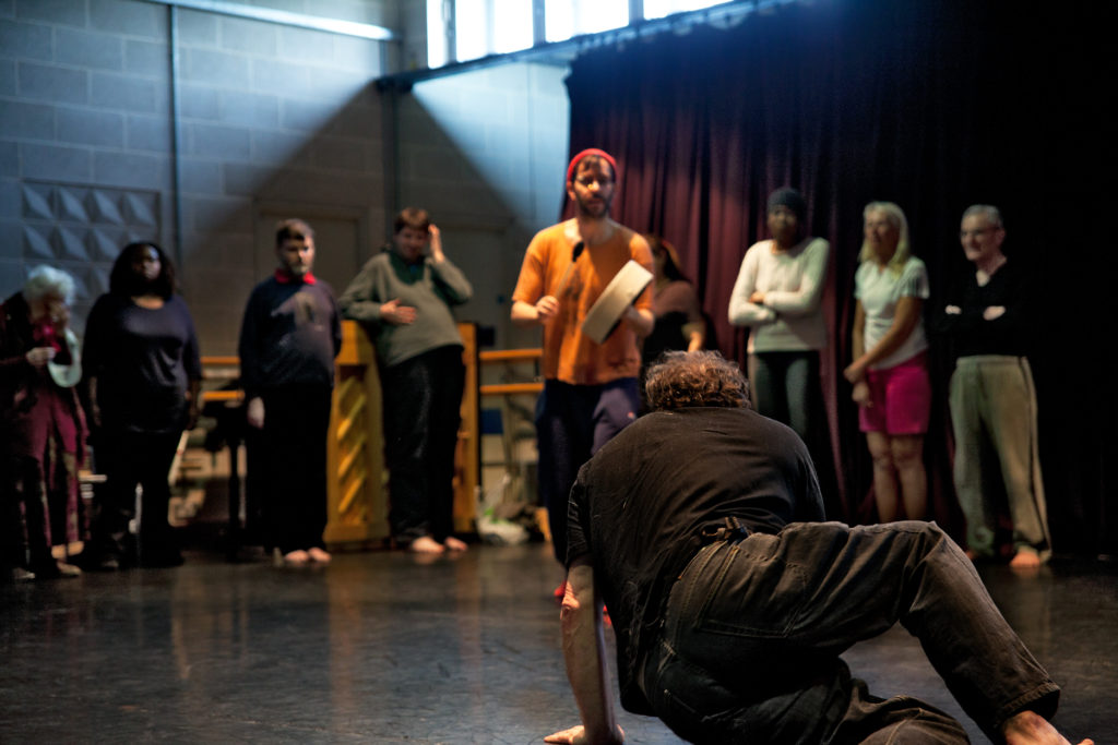 A group of people standing in a circle in a rehearsal room. Foreground, a man crouching on the ground with his back to the camera, facing the other participants.