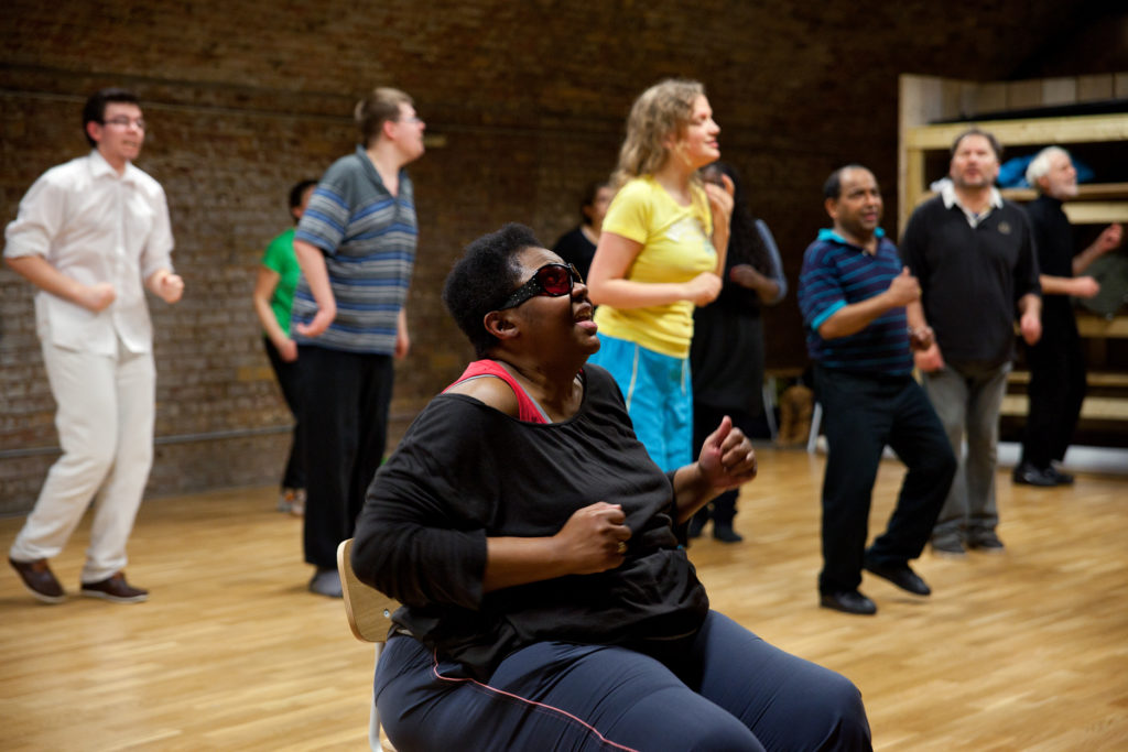 A black woman in sunglasses sits smiling as she joins in with a Zumba class from a chair