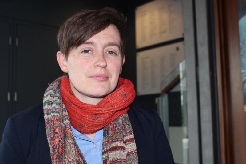 Hannah looking to camera with a neutral expression. She wears a navy blazer over a pale blue shirt, and a red scarf, parts of which have a paisley-like pattern. Hannah's hair is brown and cropped with a fringe.